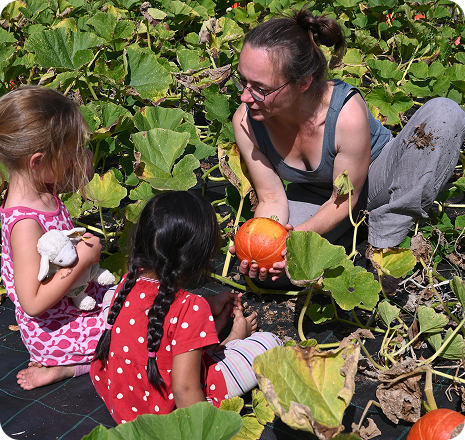Person mit Kürbis in er Hand auf dem Feld spricht mit zwei Kindern im Garten hockend
