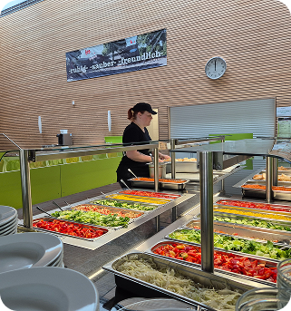 Eine Essensausgabe mit frischem Gemüse in der Auslage und einer Person die bedient dahinter